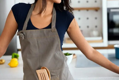 A person wearing a gray apron with wooden kitchen utensils in the pocket, standing in a kitchen with a white countertop, a blue pot on the stove, and fresh ingredients like a lemon and green bell pepper nearby.
