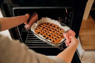 Hands in pink checkered oven mitts place a glass baking dish with a lattice‑topped pie on parchment paper into a dark oven, with a wooden countertop and paper towel roll visible nearby.