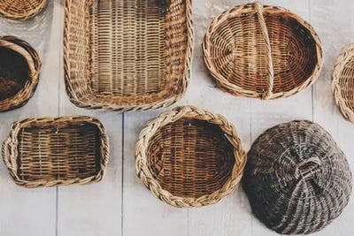Assorted woven baskets in natural tones arranged on a light wooden surface—round, oval, and rectangular shapes with varied weave patterns, including a dark circular basket and handled pieces.