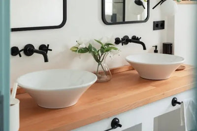 A modern bathroom vanity with a wooden countertop, two white vessel sinks, matte black wall-mounted faucets, and framed mirrors. Decor includes white lilies in a vase and sleek black accessories.