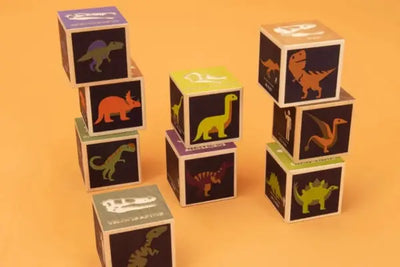 Wooden blocks with colorful dinosaur illustrations and names, arranged on an orange background. Features silhouettes, skulls, and species like T-Rex, Velociraptor, and Brachiosaurus.