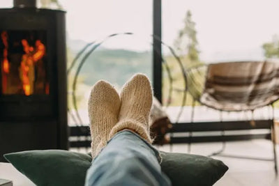 Cozy scene with feet in knitted socks resting on a green pillow, light blue jeans, a warm wood-burning stove, and a scenic outdoor view of greenery and mountains through a glass window. A striped blanket drapes over a chair, adding to the relaxed vibe.
