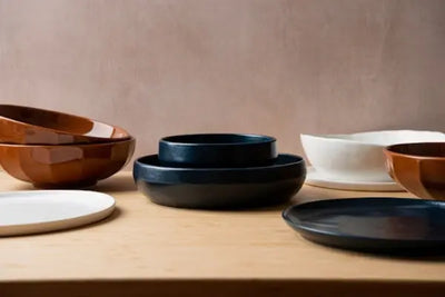 Assorted ceramic bowls and plates on a light wooden surface against a warm reddish-brown backdrop, featuring faceted brown bowls, matte navy pieces, and an organic white bowl on a saucer.
