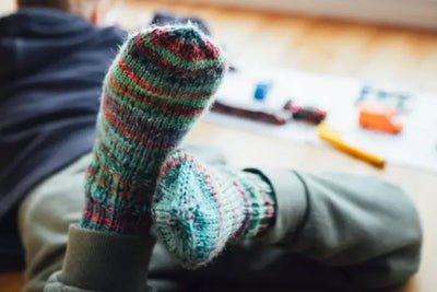 Close-up of feet in colorful, hand-knit striped socks resting on a light wood floor. Blurry background shows scattered kids’ toys and paper, creating a cozy, playful indoor scene.
