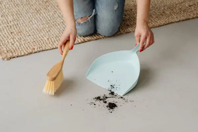 Kneeling person in ripped jeans sweeping a small pile of dirt into a light blue dustpan with a wooden hand broom on a light floor; natural fiber rug visible in the background.