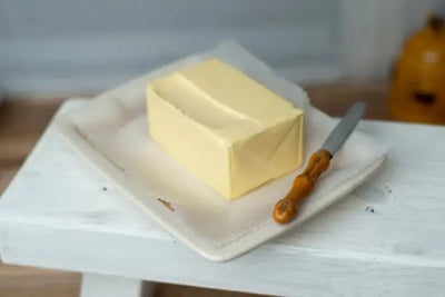 Square ceramic dish on a small white stool holding a partially unwrapped block of butter and a butter knife with a carved wooden handle; soft, rustic kitchen setting with blurred background.