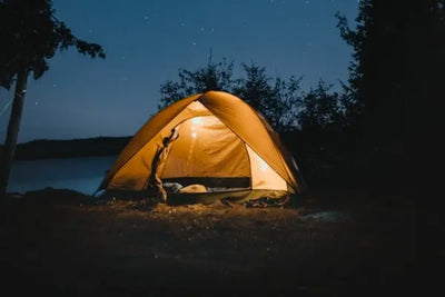 A glowing orange tent under a starry night sky, surrounded by dark tree silhouettes and near a calm body of water. The warm tent light contrasts beautifully with the serene, natural nighttime setting.