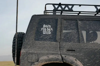 Mud-covered off-road vehicle with a spare tire and roof rack, parked in a natural setting. The rear window displays the phrase 'Born to be Wild' in a bold, stylized font, emphasizing adventure and outdoor exploration.