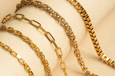 Close-up of gold chain bracelets on a textured neutral background, featuring various styles like rectangular links, twisted rope, elongated ovals, and flat box designs. The polished finish highlights their luxurious and elegant craftsmanship.