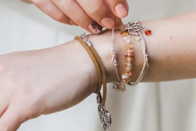 Close-up of a woman adjusting layered charm bracelets featuring amber stones, a silver tree pendant, delicate chain accents, and suede cords on her wrist in soft natural light against a neutral background.