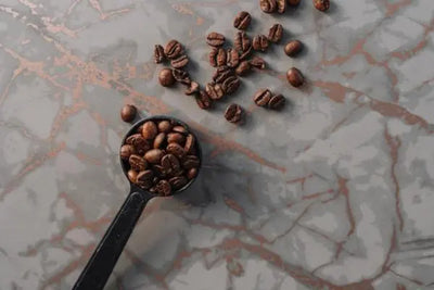 A black scoop filled with coffee beans, placed on a marbled surface.