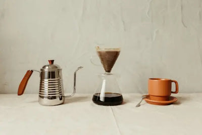 A coffee setup with a silver kettle, a glass coffee maker with brewed coffee, and a brown cup on a saucer.