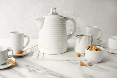 A white teapot on a marble surface, surrounded by cups and sugar cubes in bowls.