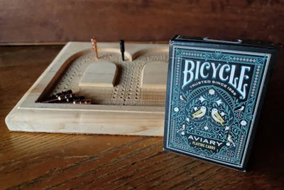 A Bicycle Aviary playing cards box next to a wooden cribbage board and pegs.