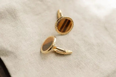 Gold cufflinks with polished Tiger's Eye stones resting on light beige fabric. The stones feature rich brown and golden bands, highlighted by soft lighting, showcasing elegance and craftsmanship.