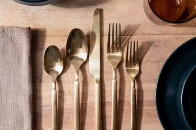 Top-down flat lay on light wood: five gold utensils aligned (small spoon, large spoon, knife, dinner fork, salad fork), beige linen napkin left, dark plate right, amber glass top-right. Minimal, refined.