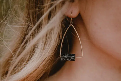 A close-up of a woman's ear showing a large geometric earring with black stone and golden frame.