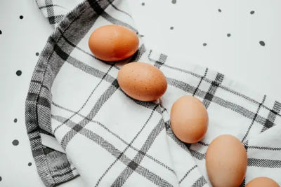 Brown eggs arranged on a white and gray plaid cloth over a white surface with scattered black polka dots; minimalist flat lay with a cozy, kitchen-inspired aesthetic.