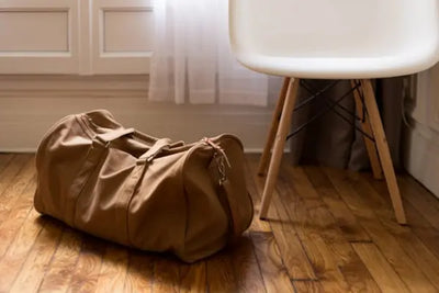 A brown duffel bag on wooden floor next to a white chair. 