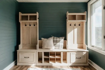 A light wood mudroom unit with lockers, a bench, and cubbies against a dark teal shiplap wall. Decorated with pillows, it’s illuminated by natural light from a large window, creating a cozy, organized space.