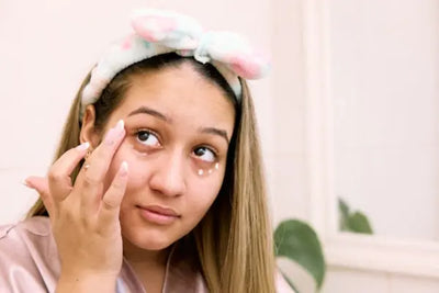 A woman wearing a pink nightgown applying American made eye care and creams on her face.