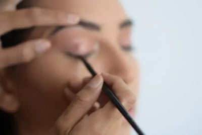 A close-up of black eyeliner being applied to a person's upper lash line with a fine brush. One hand holds the brush, while another gently stretches the eyelid. Soft lighting highlights the precision.