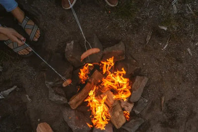 Two people cooking hot dogs over a campfire surrounded by rocks. 