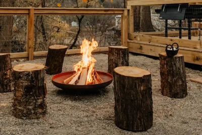 A round fire pit with burning logs surrounded by wooden stump seats on gravel.