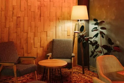 Cozy lounge with a textured wooden accent wall, gray armchairs, a round coffee table, a tripod floor lamp with a conical shade, and a potted plant. Warm lighting and earthy tones create a relaxing vibe.