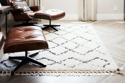 Cozy living space with a white shag rug featuring black geometric lines, brown leather lounge chairs and ottomans, and light herringbone wood floors near a window with soft curtains.
