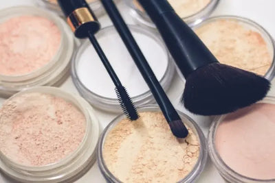 A close-up of loose powders in clear jars, makeup brushes, and a mascara wand on a soft, neutral background. The powders range from peach to beige, with soft lighting highlighting the beauty tools.