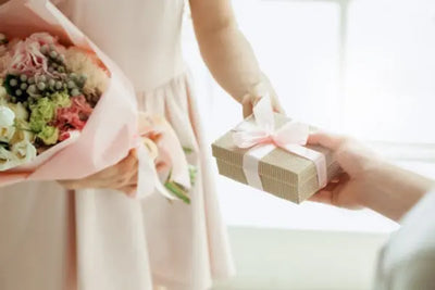 A pastel-toned scene of a gift exchange, featuring a bouquet of pink and white flowers and a textured gift box with a white ribbon, symbolizing a celebratory moment.