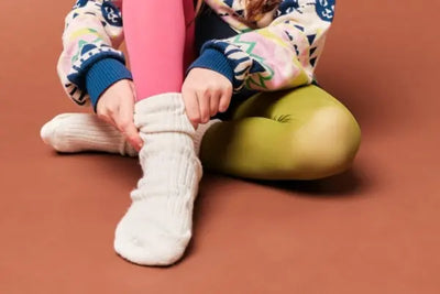 Close-up of a seated person adjusting a white ribbed sock. One leg wears hot pink tights, the other lime green, paired with a colorful patterned sweater against a reddish-brown background.