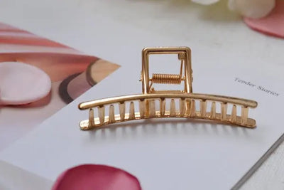 Gold hair clip with a rectangular clasp on an open magazine page showing 'Tender Stories.' Surrounded by soft pink and white petals, the blurred background highlights the clip's polished, elegant design.