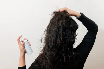 Back view of a person with long, wavy dark hair, holding a white spray bottle in one hand while styling their hair with the other. Neutral background highlights the hair care action.