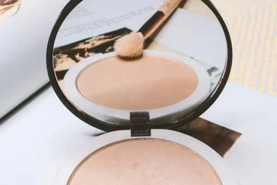 An open compact of shimmery champagne highlighter with a built-in mirror reflecting a makeup brush and magazine pages. The compact rests on a light surface with soft lighting and a striped background.