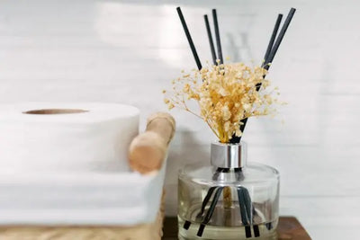 A glass diffuser with black reeds and dried yellow flowers next to rolls of toilet paper. 