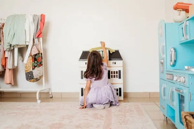Child in a purple dress plays with a white dollhouse in a bright room. A clothing rack with colorful outfits and a blue toy kitchen surround the scene, creating a cozy and imaginative play area.