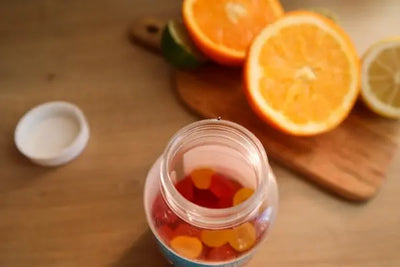 A jar of colorful red and orange gummy candies sits on a wooden surface with citrus fruits, including oranges, lime, and lemon, blurred in the background. The scene highlights fruity, natural flavors.