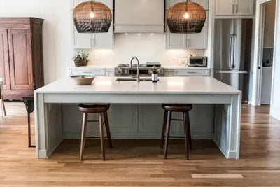 A modern kitchen with a large island, light countertops, gray cabinets, stainless steel appliances, wooden bar stools, and woven pendant lights. Warm hardwood floors and natural accents complete the look.