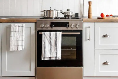 Stainless steel oven with striped towel on handle, flanked by light cabinets and wood countertops; pots on stove, tomatoes and knife on cutting board, white tile backsplash in a bright modern kitchen.