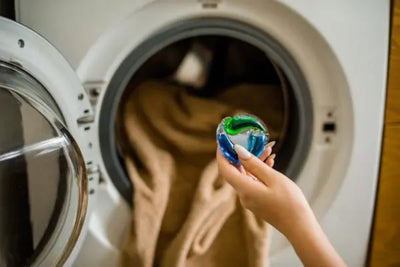 Hand holding a blue-green laundry detergent pod in front of an open front-loading washer with tan clothes inside; close-up focus on the pod, background slightly blurred.