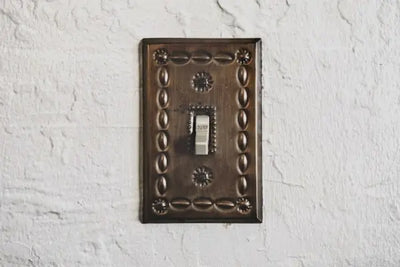 Vintage-style toggle light switch with an ornate brass faceplate featuring embossed patterns, mounted on a textured off-white wall. The switch is in the 