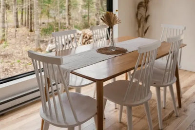 A cozy dining area featuring a wooden table with a white table runner, a glass vase of dried flowers, and six white wooden chairs. Natural light streams through a large window, highlighting the minimalist decor and natural elements.