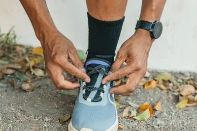 Close-up of a person tying laces on a light blue running shoe outdoors. Black athletic socks and a black smartwatch are visible. Fallen leaves on a dirt path suggest a fall workout or run in progress.