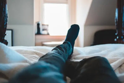 First-person view of someone relaxing in bed, legs in blue jeans and dark socks stretched toward a bright window. Soft, rumpled cream bedding and wooden bedposts create a cozy, tranquil bedroom scene.