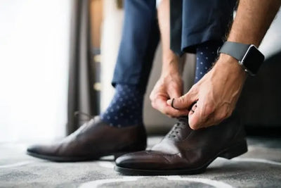 Close-up of a person tying brown leather dress shoes on a patterned carpet. Navy trousers and dark blue polka-dot socks are visible, along with a smartwatch, capturing a polished, getting-ready moment.