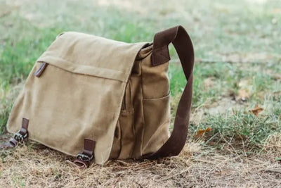 A brown canvas messenger bag resting on grassy ground with a brown strap.
