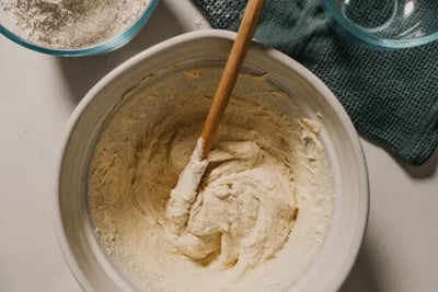 Overhead view of a large ceramic mixing bowl with beige batter and a wooden spatula, beside a glass bowl of flour and another empty glass bowl on a dark teal textured cloth on a light countertop.