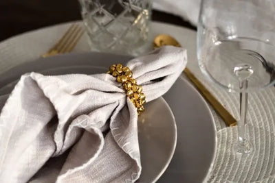 Close-up of a place setting with a light linen napkin held by a gold nugget-style napkin ring on a matte gray plate, paired with gold flatware and clear glassware on a textured light placemat.
